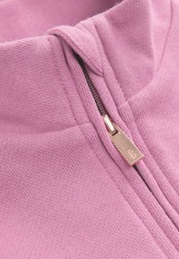 Sudadera con cremallera de color lila claro, que presenta un alto cuello y una cremallera metálica con un sutil acento de marca. Tejido suave y texturizado.