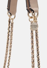 Cintura in pelle beige e nera con accenti in catena dorata, dotata di un'etichetta circolare con la scritta "GUESS EST. 1981" attaccata vicino al meccanismo.