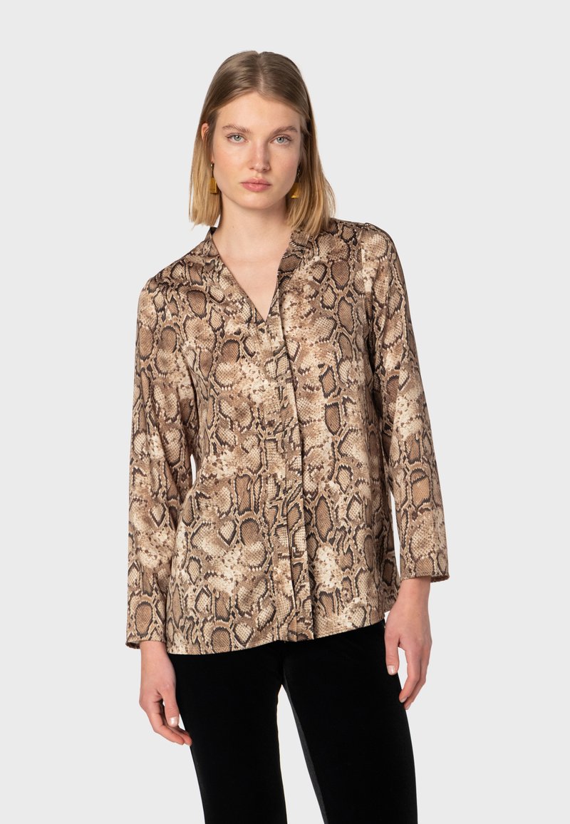 Blusa a maniche lunghe con stampa serpente in tonalità beige e marrone. Caratterizzata da scollo a V, tessuto morbido e vestibilità ampia, abbinata a pantaloni neri.