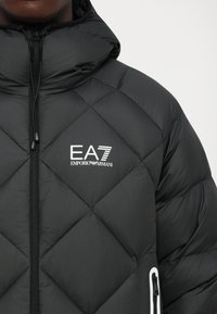 Jachetă neagră matlasată cu glugă, fermoar frontal, logo alb EA7 Emporio Armani pe piept și detalii cu buzunar cu fermoar pe braț.