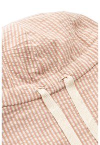 Prugasta kapa od mekanog bež i bijelog platna s teksturiranim završetkom. Pruža mogućnost podešavanja pomoću vrpce na dnu.