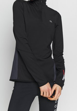 Henkilö, jolla on musta pitkähihainen urheilupaita, jossa on neljännesvetoketju, ja mustat legginsit valkoisilla logokuvilla reiden kohdalla, yksivärisen taustan edessä.