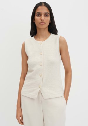 Frau mit langen dunklen Haaren, die eine ärmellose, elfenbeinfarbene, knöpfbare, texturierte Weste und passende helle Hose trägt, steht vor einem einfarbigen Hintergrund.