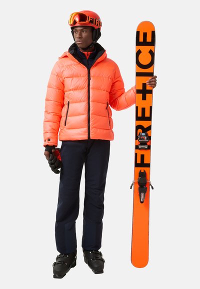 Jasnopomarańczowa, pikowana kurtka męska, granatowe spodnie, czarne buty narciarskie, pomarańczowe narty z czarnymi napisami oraz czerwony kask z goglami.