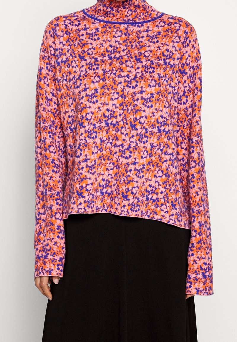 Blouse à manches longues col roulé avec imprimé floral abstrait orange, rose et violet portée avec une jupe noire.