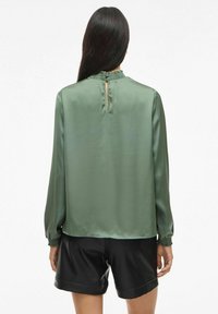 Femme aux longs cheveux bruns portant un blouse en satin vert avec un dos à ouverture en bouton et un short en cuir noir, de dos face à l'appareil photo.