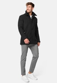 Abrigo de lana negro con cuello alto, que presenta tres botones y bolsillos laterales, combinado con pantalones gris ajustados y zapatillas blancas.