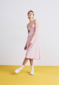 Rosa midiklänning med spagettiband, samlad byst och knytdetalj. Matchad med vita sneakers och ett pärlhalsband. Gul bakgrund.