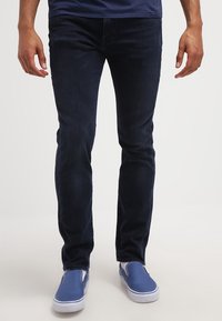 Jeans slim-fit in denim blu scuro con una trama liscia e design minimalista. Abbinati a scarpe slip-on blu con suole bianche.