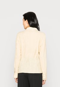 Camisa de manga longa bege com acabamento texturizado, apresenta um decote com gola e punhos abotoados. Corte ajustado, design minimalista.