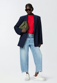 Blazer overdimensionato blu navy, camicia rossa aderente, jeans a gamba larga azzurri chiari con orlo sfilacciato, borsa verde e sneaker bianche con dettagli grigi.