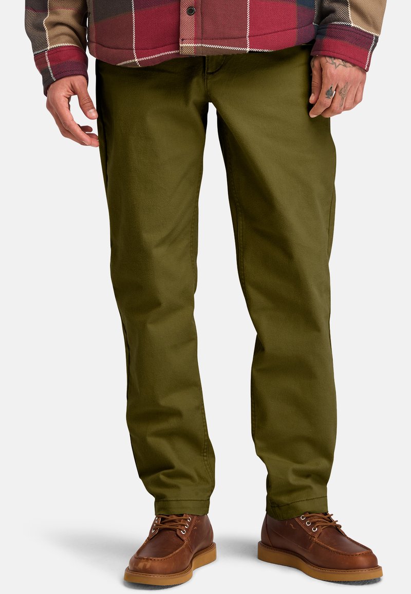 Pantaloni in cotone verde oliva con taglio dritto, tasche laterali e una texture liscia; abbinati a scarpe in pelle marrone.