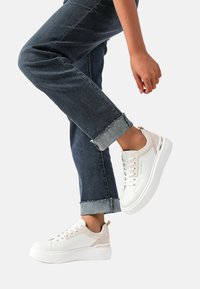 Sneakers bianchi con accenti beige, una parte superiore testurizzata e una suola spessa bianca; indossati con jeans in denim scuro arrotolati.