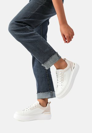 Sneakers bianchi con accenti beige, una parte superiore testurizzata e una suola spessa bianca; indossati con jeans in denim scuro arrotolati.