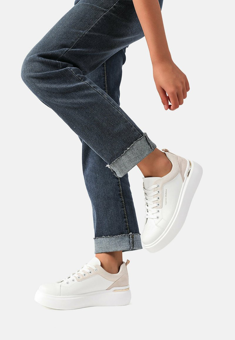 Sneakers bianchi con accenti beige, una parte superiore testurizzata e una suola spessa bianca; indossati con jeans in denim scuro arrotolati.