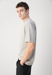 T-shirt gris clair à manches courtes en mélange de coton, avec un col rond et une coupe décontractée, associé à un pantalon noir.