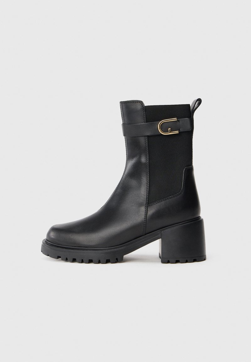 Bota de cano curto em pele preta com salto robusto, painel lateral elástico, laço traseiro e tira decorativa com fivela dourada em fundo neutro.