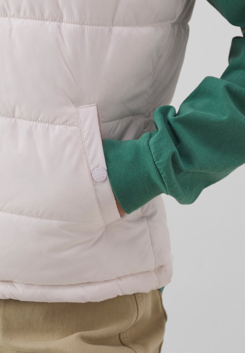 Leichte pinke Steppweste mit Druckknopf-Taschen-Detail, getragen über einem grünen Langarmshirt. Die Textur wirkt glatt und leicht glänzend.