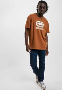 Brun bomull t-shirt med korte ærmer og et frontgrafik; parret med mørkeblå jeans og grå-hvide sneakers med accenter.
