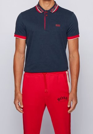Granatowa koszulka polo z paskowanym kołnierzykiem, z czerwonymi akcentami i trzema guzikami. W zestawie z czerwonymi dresami, które mają logo i sznurek.