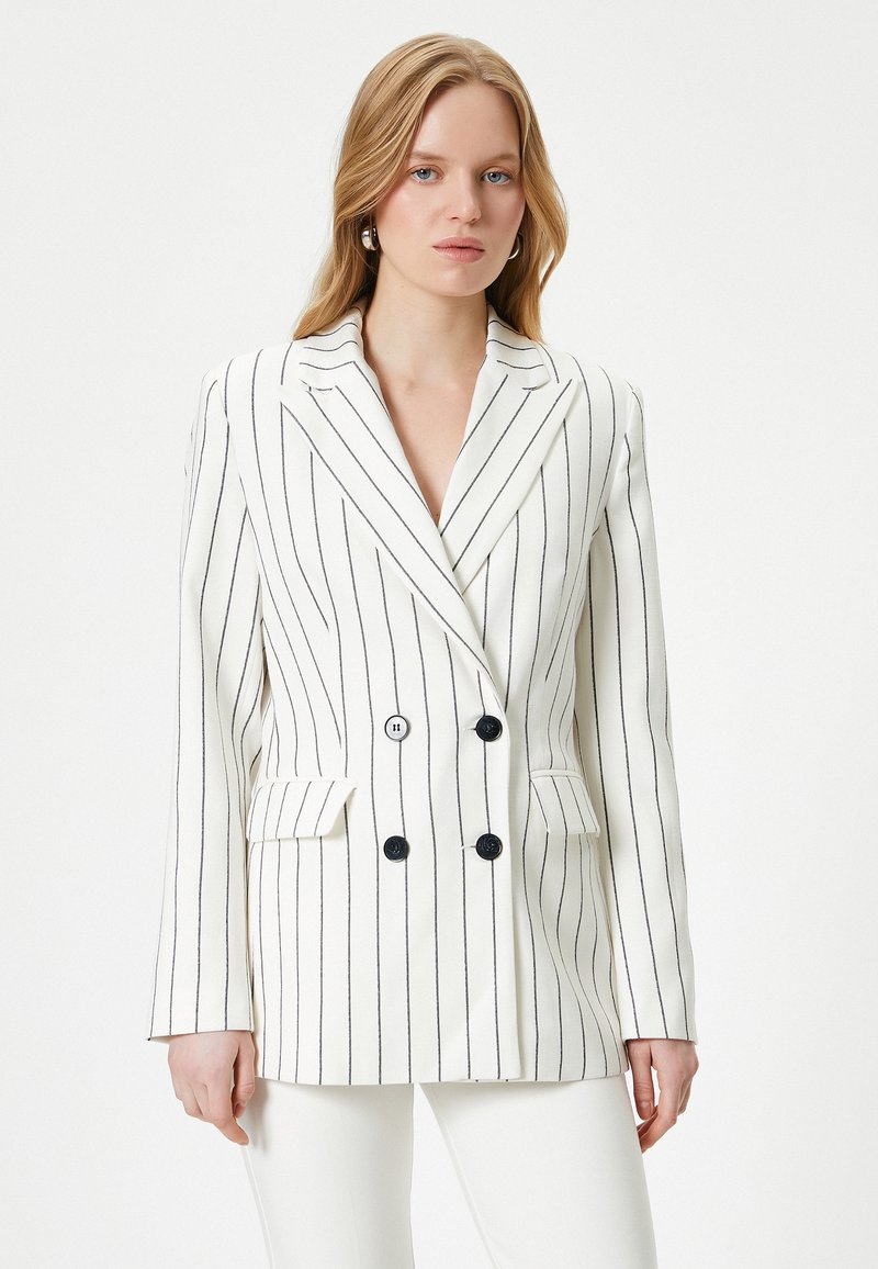 Blazer de doble botonadura en blanco con rayas negras, que presenta una solapa de pico, dos bolsillos frontales y botones negros.