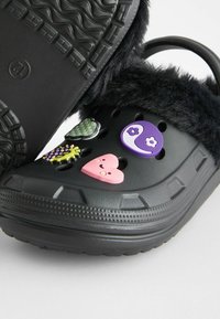 Crocs en fourrure noire avec semelle en caoutchouc, ornés de pin's décoratifs colorés incluant des cœurs, un yin-yang et des motifs floraux.