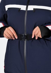 Námořnická venkovní bunda s bílými a červenými pruhy, černým zipem a nastavitelným pásem. Má dlouhé rukávy s černými manžetami a kapsami na zip.