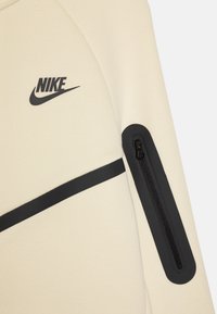Svijetlosmeđi Nike dukserica s crnim logotipom, s patentnim zatvaračem na rukavu i crnom akcentnom prugom preko prednje strane.