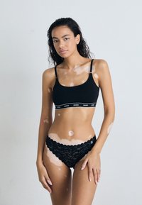 Soutien-gorge de sport noir avec bande logo ; culottes en dentelle noire à bords festonnés ; texture lisse sur l'ensemble ; le mannequin se tient les mains sur les hanches.