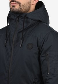 Veste bleu marine foncé avec un col montant, une capuche ajustable et des poches latérales zippées. Comprend un logo rond et des détails en tissu texturé.