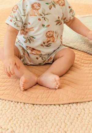 Bébé assis sur un tapis beige texturé, portant une combinaison blanche ornée d'imprimés de lion brun et de feuilles vertes, avec les bras et les jambes visibles.