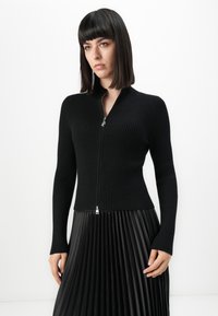 Woman with straight dark hair wearing a fitted black ribbed zip-up sweater and black pleated skirt, standing against a plain white background.