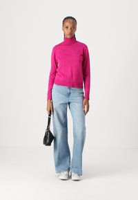 Strakke coltrui in felroze, gecombineerd met lichtblauwe wijde jeans. Model houdt een kleine zwarte handtas vast en draagt witte sneakers.