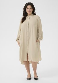 Beige knoopjurk met lange mouwen, zijzakken en een high-low zoom, gemaakt van lichtgewicht stof. G gedragen met zwarte puntige schoenen.
