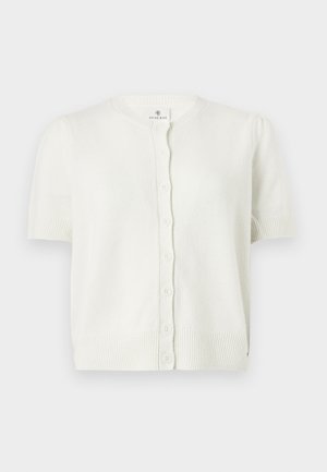 Femme portant un pull blanc à manches courtes boutonné et un pantalon large bleu marine, debout avec une main dans la poche devant un fond uni.