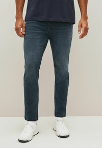 Tamno plave traperice slim fit kroja, s klasičnim dizajnom s pet džepova i suptilnim izblijedjenjem. Nošene uz bijele tenisice.