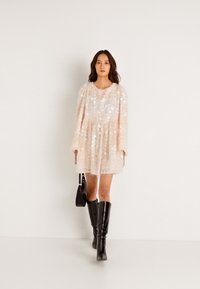 Ljusrosa klänning med långa ärmar, strukturerad tyg, lagerdesign och rund halsringning; matchad med knähöga svarta stövlar och en svart väska.