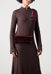 Sac bandoulière en cuir verni rouge de forme rectangulaire, avec rabat avant et sangle réglable, assorti à une robe marron côtelée.