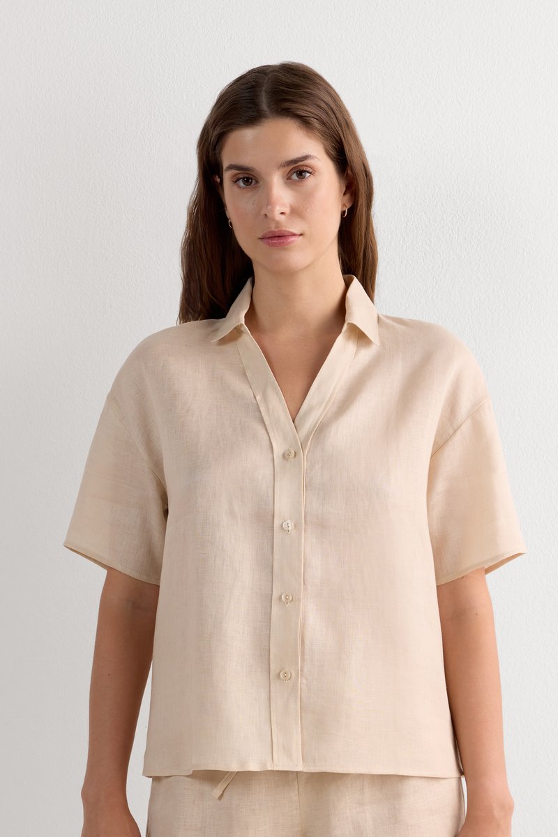 Vrouw met bruin haar, gekleed in een beige korte-mouw blouse met knopen en bijpassende broek, staand tegen een vlakke witte muur.