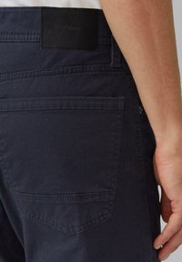 Dunkelblaue Hose mit strukturierter Stoffqualität, erkennbarer Gesäßtasche und einem mattschwarzen Markenlabel in der Nähe des Bunds.