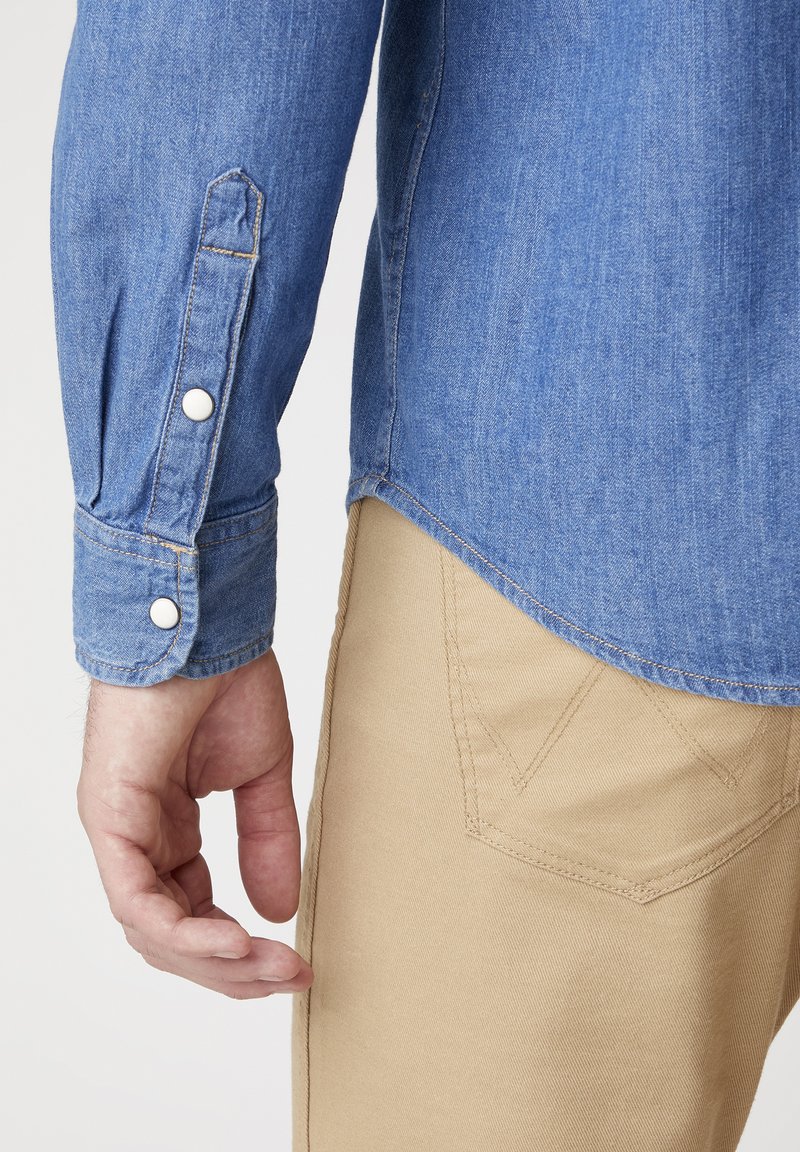 Camisa de mezclilla con una textura azul, con botones blancos y puños doblados. Pantalones beige con una sutíl costura diagonal y detalles en los bolsillos.