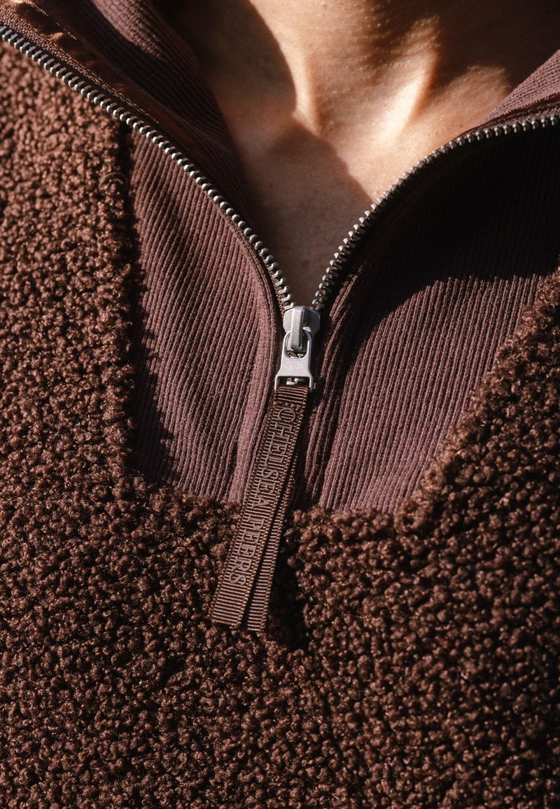 Ruskea vetoketjullinen fleece takki, jossa on teksturoitu pinta; ribattu kaulus ja vetoketju yksityiskohtia. Kangas on pehmeää ja pörröistä.