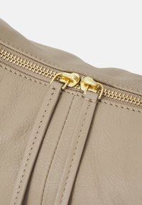 Beige läderpouch med två guldzipper högst upp. Har en sydd söm längs kanterna för extra detaljer och hållbarhet.