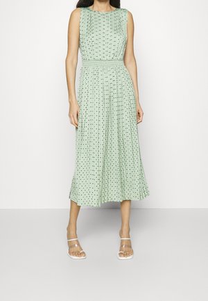 Vestido sin mangas verde menta con diseño plisado, con lunares negros, cintura fruncida y dobladillo de longitud media. Combinado con sandalias transparentes.