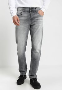 Jeans de mezclilla gris con un diseño desgastado, cinco bolsillos y cremallera. Combinados con zapatos negros de cordones, con una suela de goma gruesa.