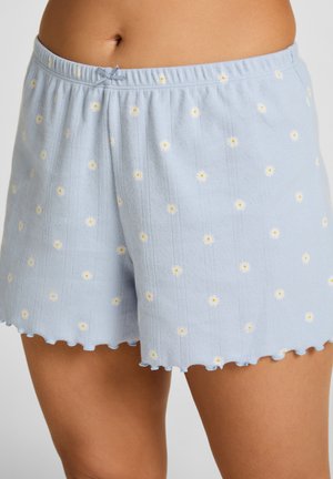 Lichtblauwe gebreide shorts met kleine witte en gele madeliefjes, een geschulpte zoom en een klein strikje op de elastische tailleband, gedragen op gebruinde huid.