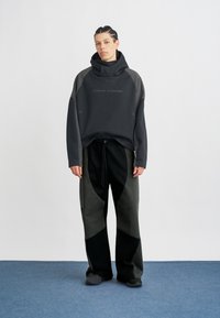 Sudadera negra con detalles en gris texturizado, con cuello alto y corte holgado. Pantalones de pierna ancha en gris y negro con parches geométricos. Calzado gris.