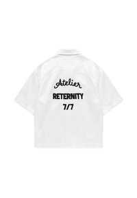 Chemise blanche à manches courtes avec texte noir au dos indiquant « Atelier RETERNITY 7/7 » en polices mixtes et gras.