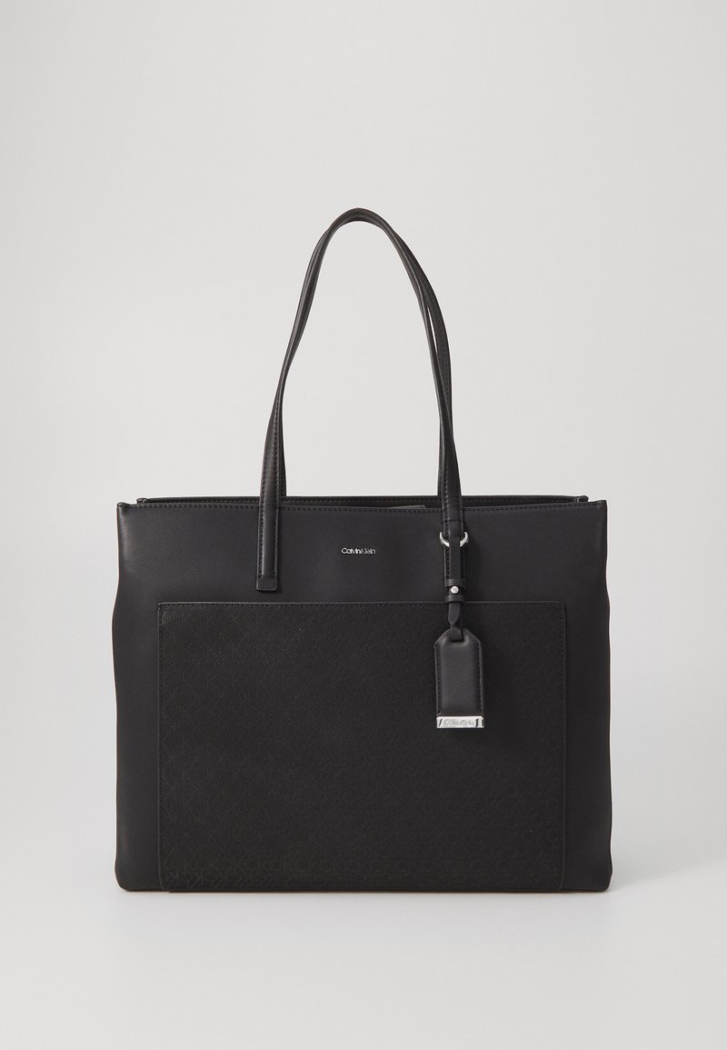 Black tote bag made of smooth leather with a patterned fabric front panel, dual handles, and a silver hardware accent. Zipper closure at the top.