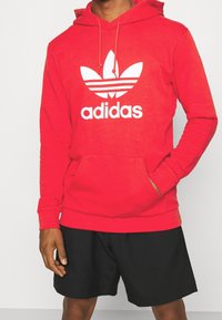 Roter Baumwoll-Hoodie mit Kängurutasche, versehen mit einem großen weißen Adidas-Logo und -Trefoil-Design. Kombiniert mit schwarzen Shorts.
